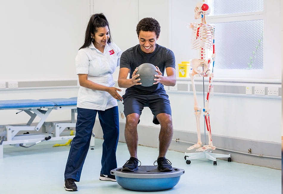 A person balances on a Bosu ball while holding a medicine ball, assisted by another person in a clinical room with a skeleton model in the background.