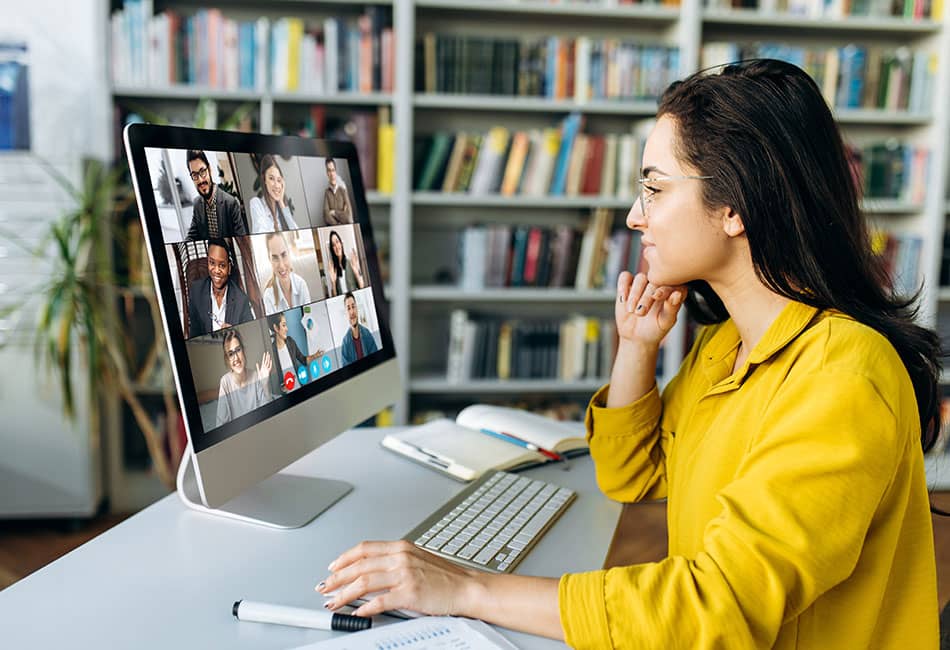 A young woman sat at a modern desk is using a computer for a group video call.