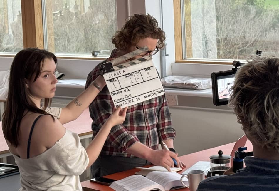 Two students working on creating a film, one is behind the camera, the other is holding a clapper board.