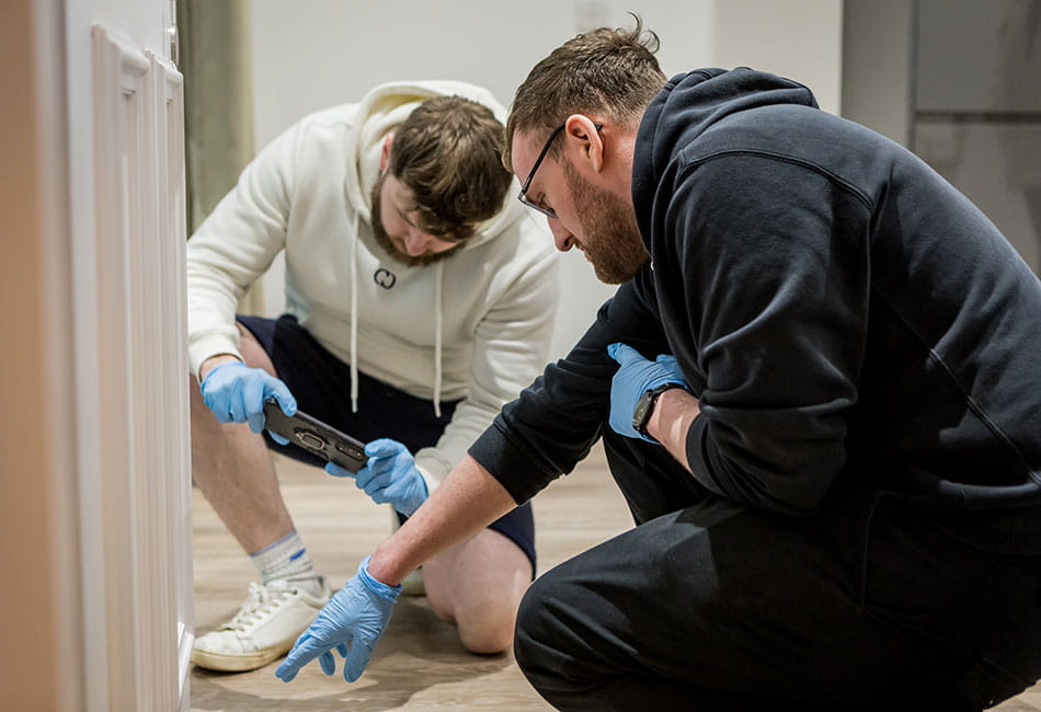 Two students wearing blue safety gloves looking closely at an interior door, one of them is taking a photo.