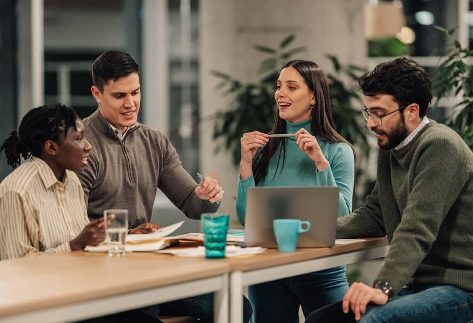 Group of people sat around a table talking with a laptop and paperwork on the table.