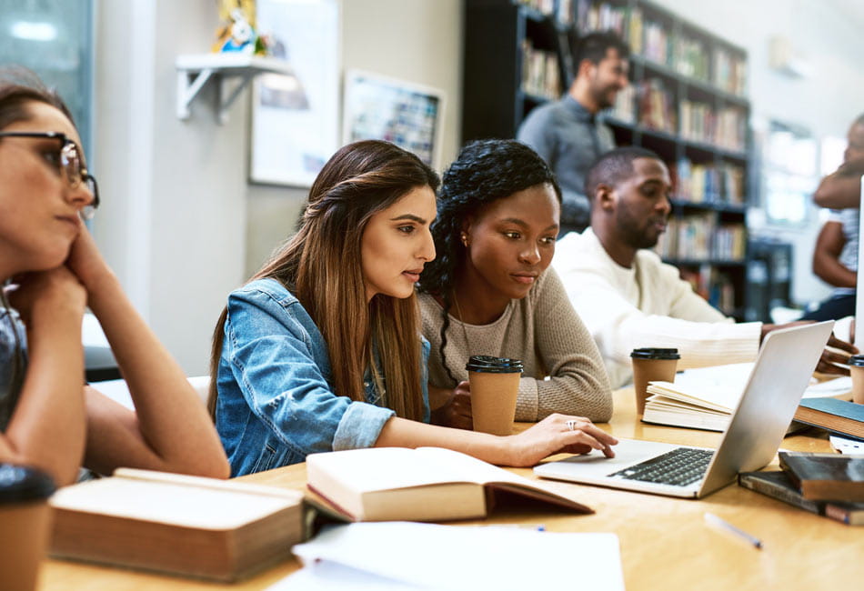 Two female students deep in focus working together at a laptop, with other students working in the background.